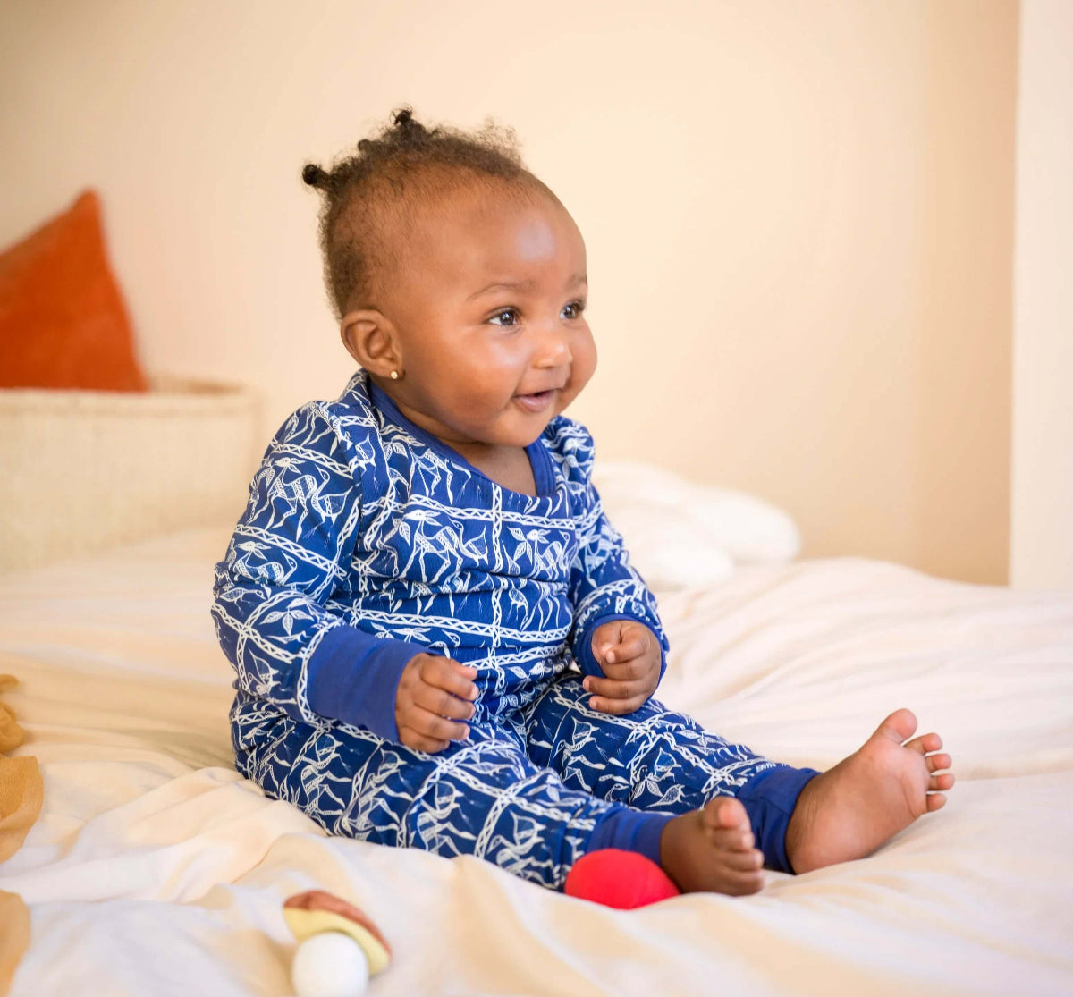 Baby wearing a blue patterned onesie/ pajama sitting on a bed with toys around.