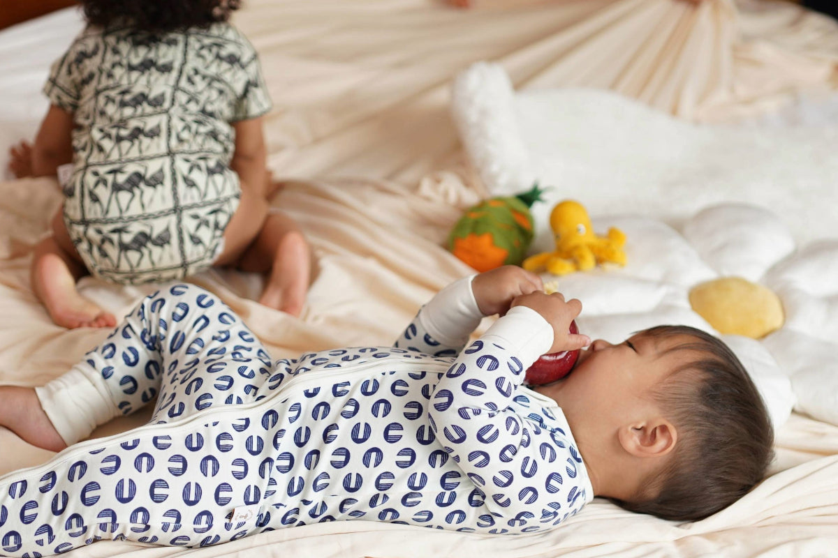 A baby lying on a cream-colored bed wearing a blue and white patterned footed ORGANIC COTTON pajama, with another baby sitting in the background wearing a green and white patterned pajama. A white plush bear toy is on the bed next to some colorful toys.