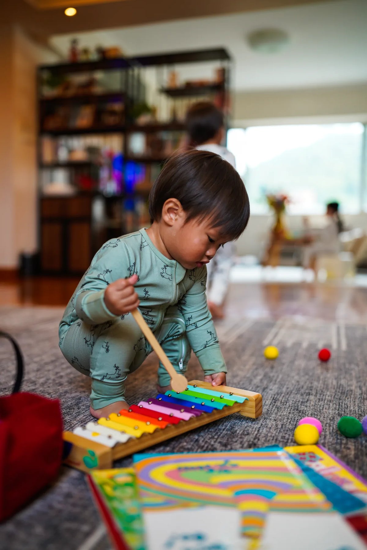 Toddler kneeling to play a toy xylophone in the KendiKids Uhuru Sage Zipper Romper — running gazelle print in soft sage