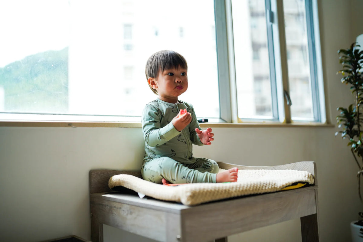 Toddler sitting by a window in the KendiKids Uhuru Sage Zipper Romper — running gazelle print in soft sage