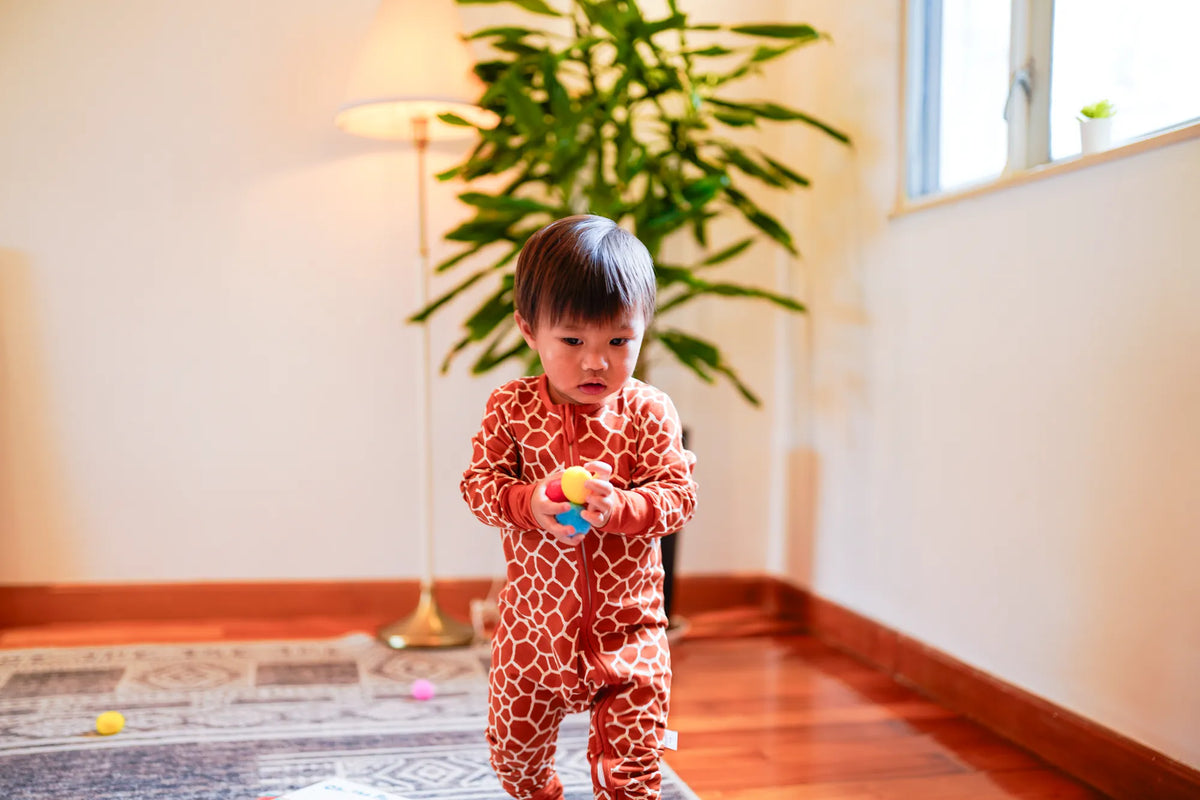Toddler standing on a rug holding a toy in the KendiKids Twiga Zipper Romper — giraffe print in burnt orange
