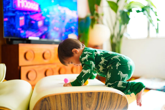 Toddler climbing over a round ottoman in the KendiKids Ndoto Zipper Romper — zebra print in white on forest green