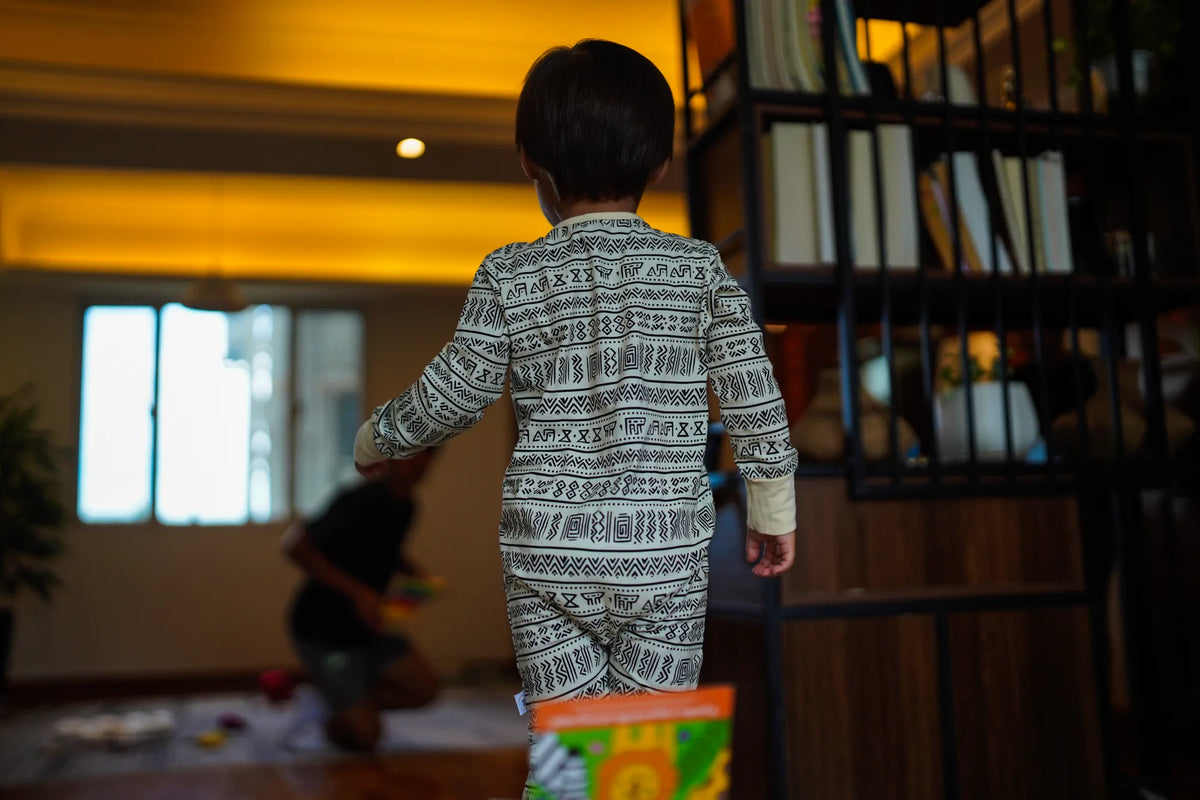 Back view of a toddler standing by a window in the KendiKids Asili Zipper Romper — mud cloth print in black on cream