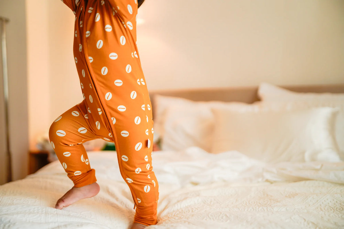 Close view of a toddler's legs on a bed in the KendiKids Baraka Zipper Romper — cowrie shell print in white on golden mustard