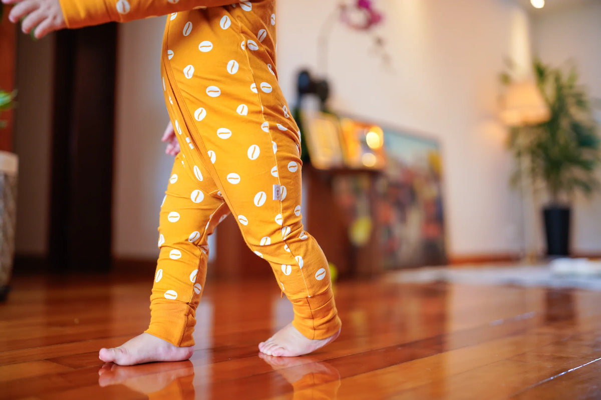 Close view of a barefoot toddler walking in the KendiKids Baraka Zipper Romper — cowrie shell print in white on golden mustard