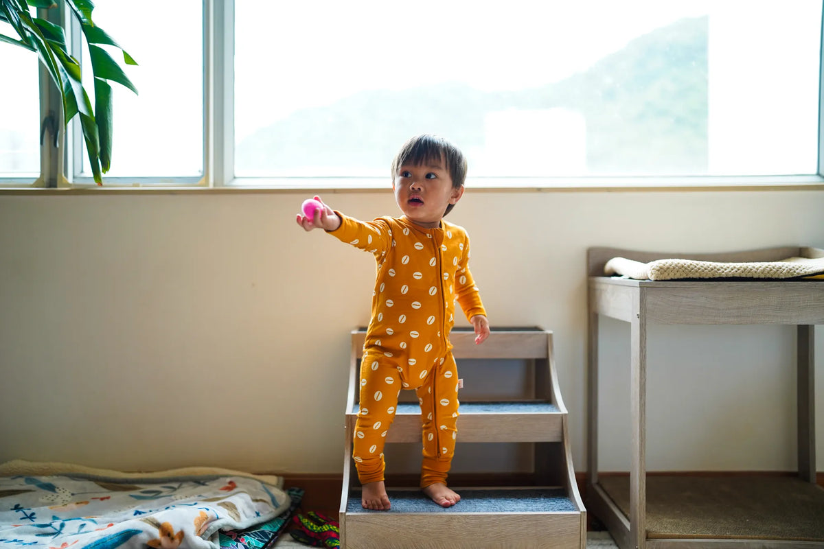 Toddler standing at a window holding a ball in the KendiKids Baraka Zipper Romper — cowrie shell print in white on golden mustard