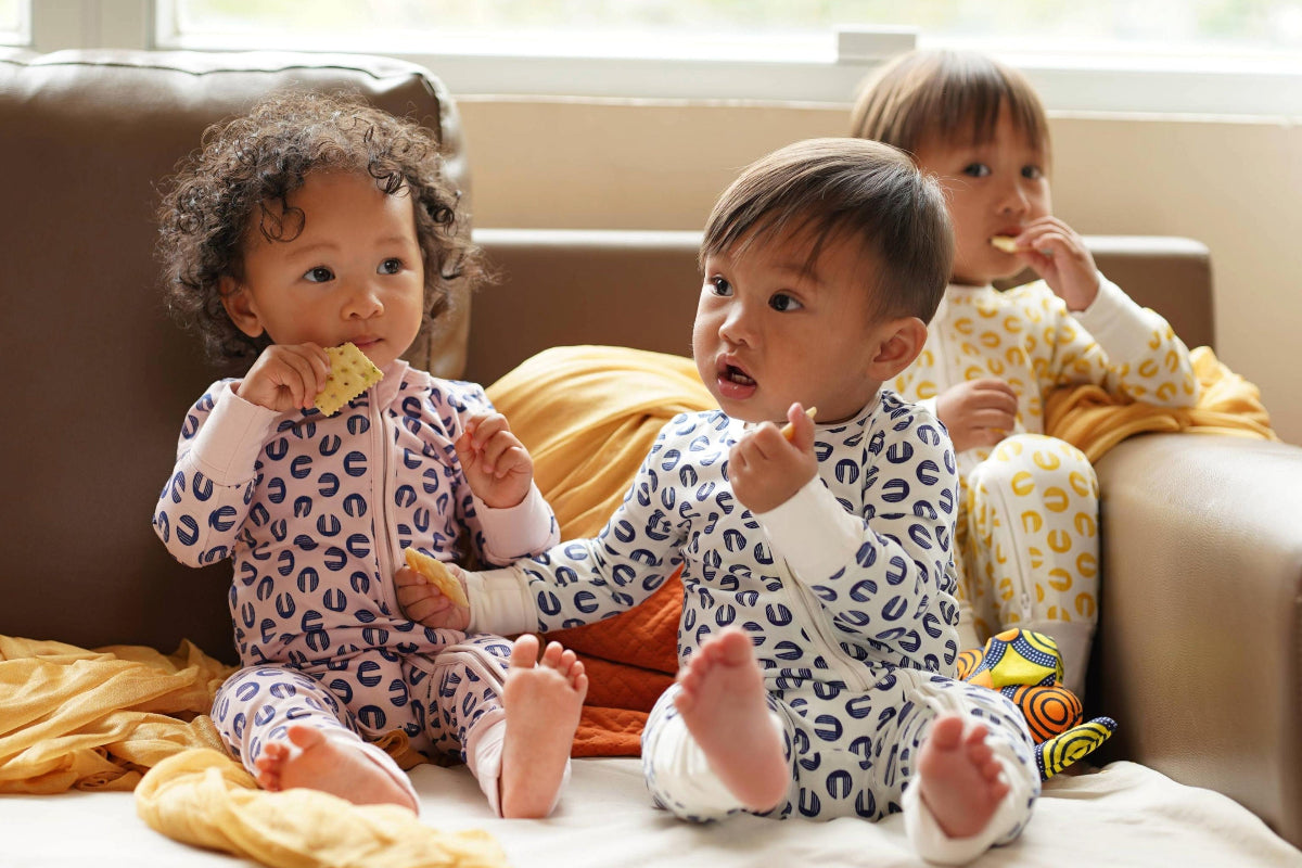 Three babies sitting on a brown leather couch wearing matching PATTERN-print one-piece ORGANIC ONESIES, ROMPER,pajamas. The pajamas feature a pattern of dark blue, purple, and yellow spots on white bases.