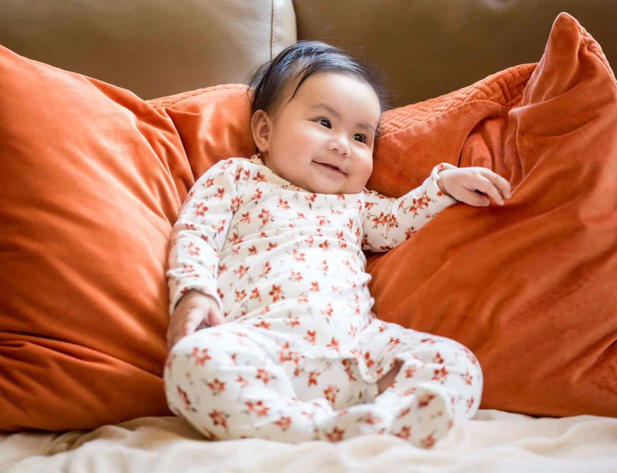 Baby in a floral onesie sitting on a couch with orange pillows