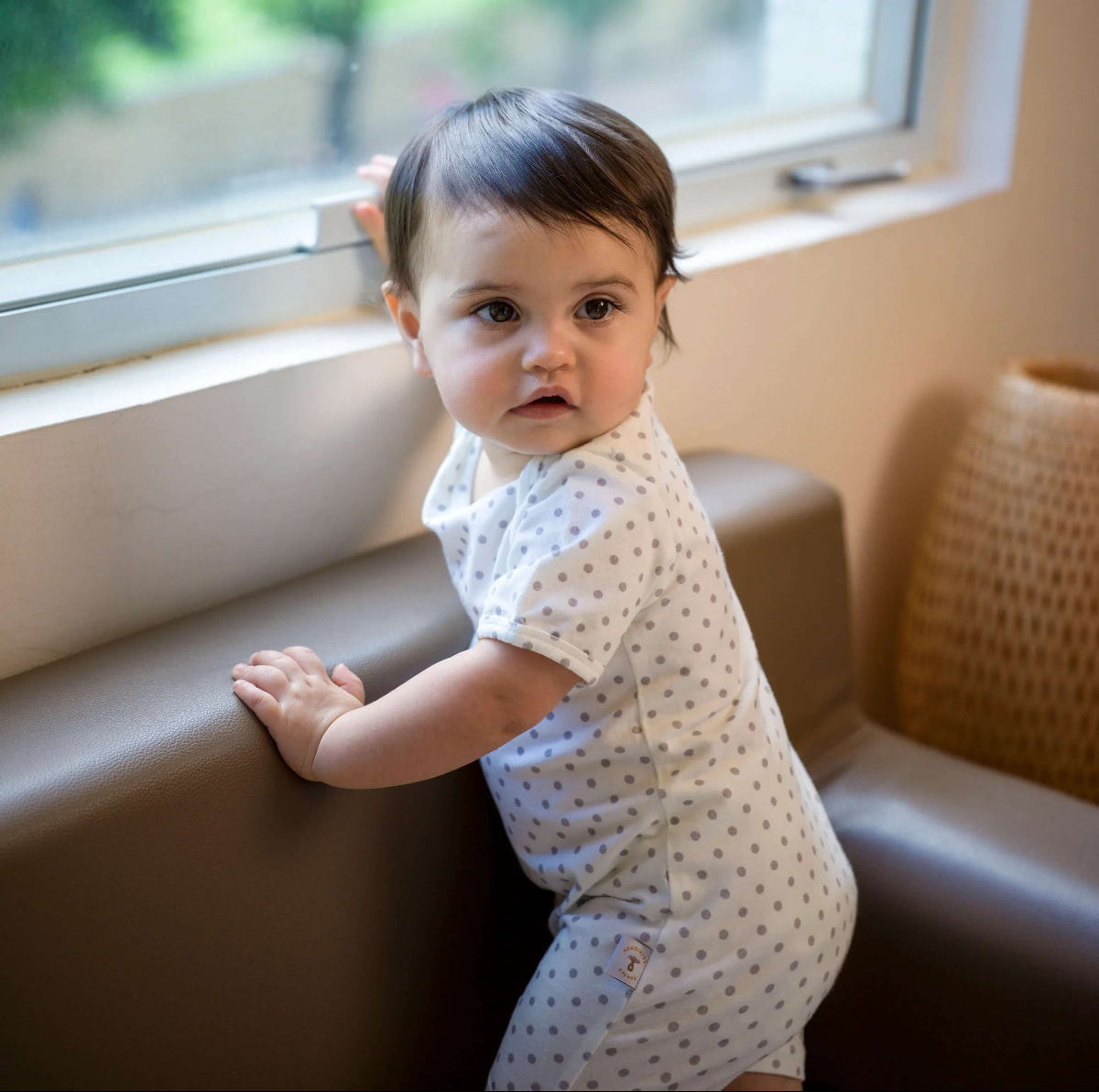 Baby in a polka dot romper standing on a chair near a window with greenery outside