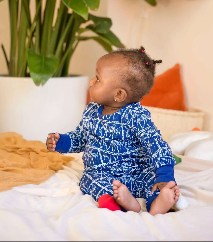 Baby in a blue onesie, pajama sitting on a bed with plants in the background