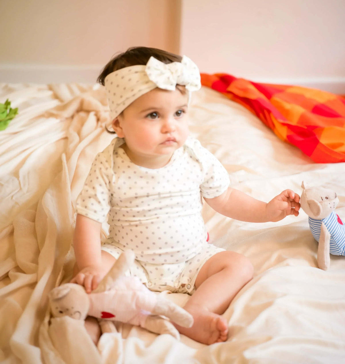 Baby sitting on a bed holding a teddy bear, wearing a white romper outfit and headband.