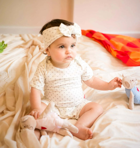 Baby sitting on a bed holding a teddy bear, wearing a white romper outfit and headband.