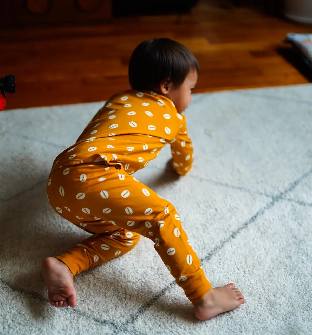 A toddler wearing a mustard yellow long-sleeve pajama set with white polka dots crawls on a white textured rug in a home setting.