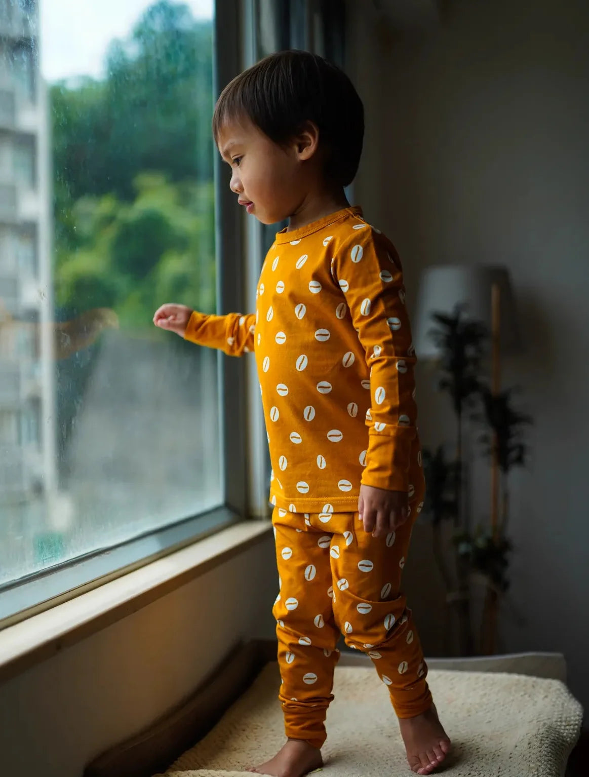 A toddler stands barefoot on a cushioned window seat indoors, wearing a bright orange long-sleeve pajama set with a white leaf pattern and looking out the window.