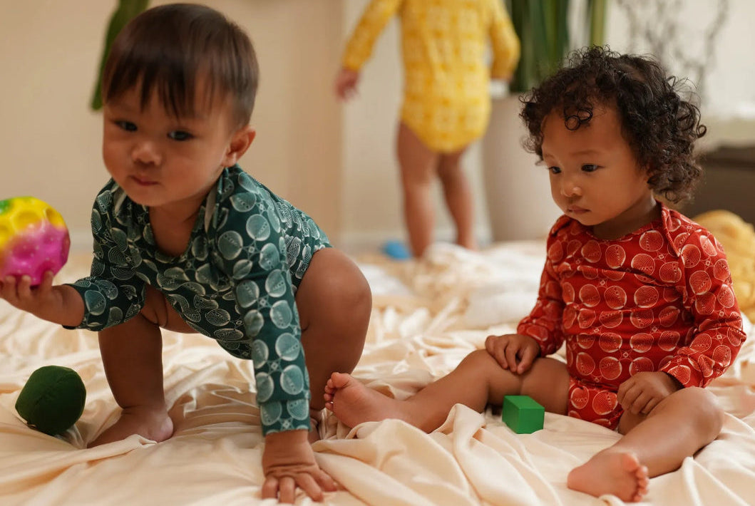 Two toddlers sitting on a cream-colored bed wearing patterned long-sleeve kendikids organic bodysuits, with one in teal and one in red-orange. A third child in a yellow bodysuit is blurred in the background.