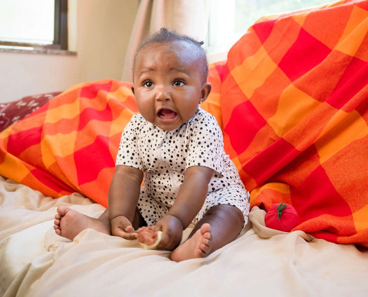 Baby sitting on a bed  wearing dotted short romperwith colorful pillows and a lamp in the background