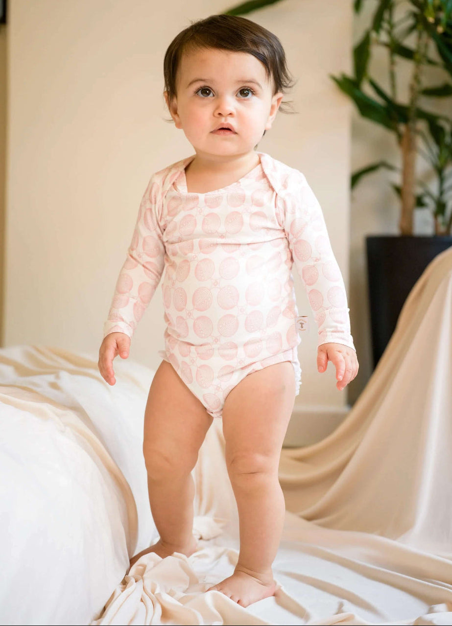 Baby in a pink patterned onesie ,bodysuit standing in a room with a plant and draped fabric.