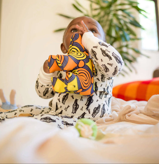 Baby wearing a colorful snap romper with zebra patterns, sitting on a bed in a room with plants and a painting.