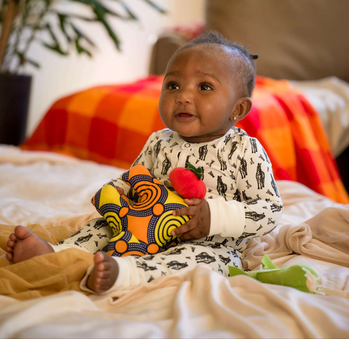Baby holding a toy on a bed with colorful blankets and a plant wearing kendikids romper